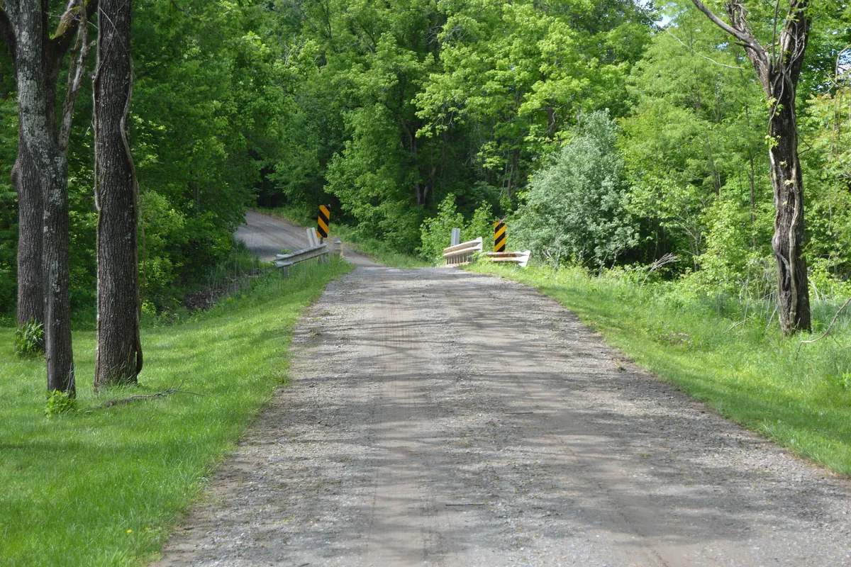 Graded gravel road running between green woods and grass toward a small bridge
