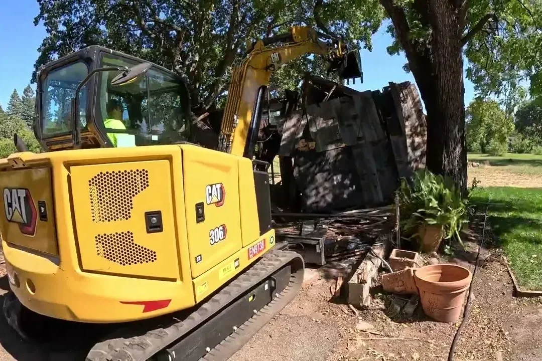Mini excavator tearing down an old wooden shed under a shade tree in a backyard