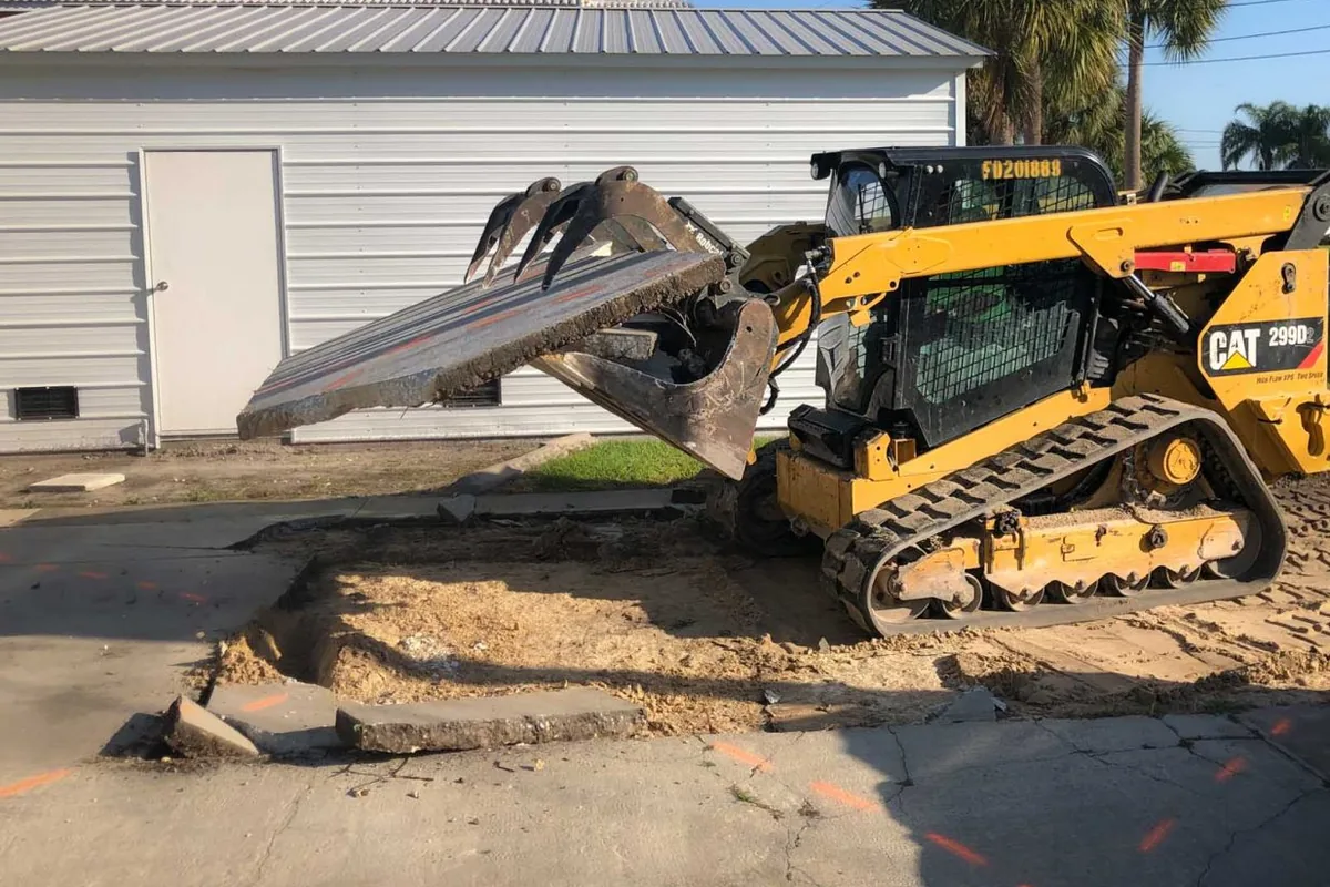 Tracked skid steer with a grapple lifting a slab of broken driveway concrete beside orange locate paint